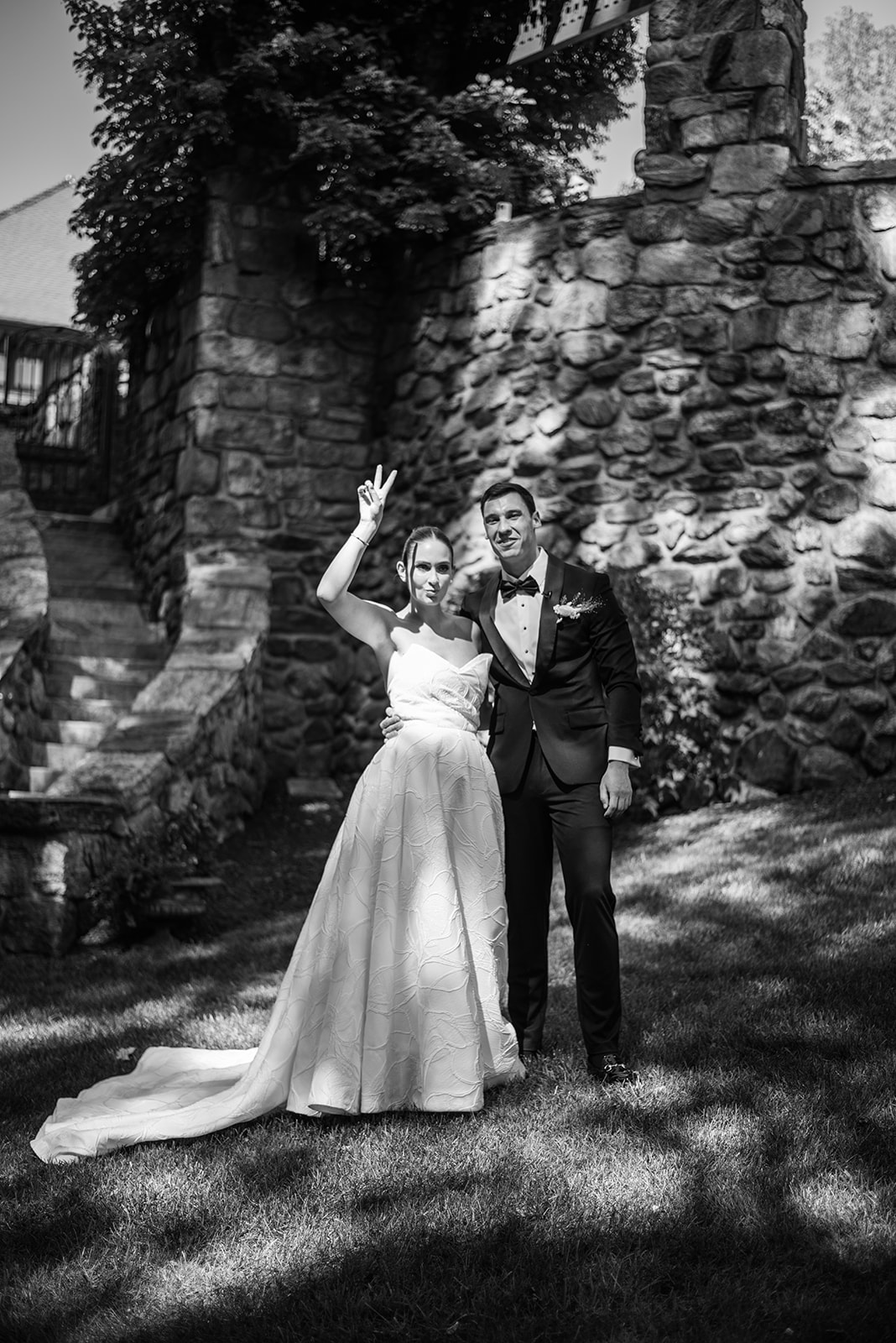 Black and white film photo of the bride flashing a peace sign beside her smiling groom.